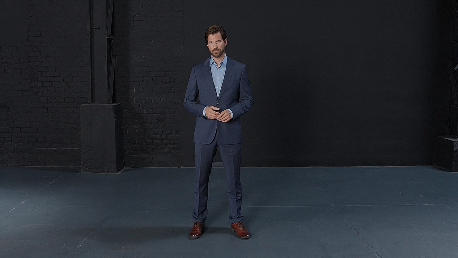 Ein professionell gekleideter Mann im Anzug blickt selbstbewusst in die Kamera. Im Hintergrund eine dunkle, moderne Loft-Wand.